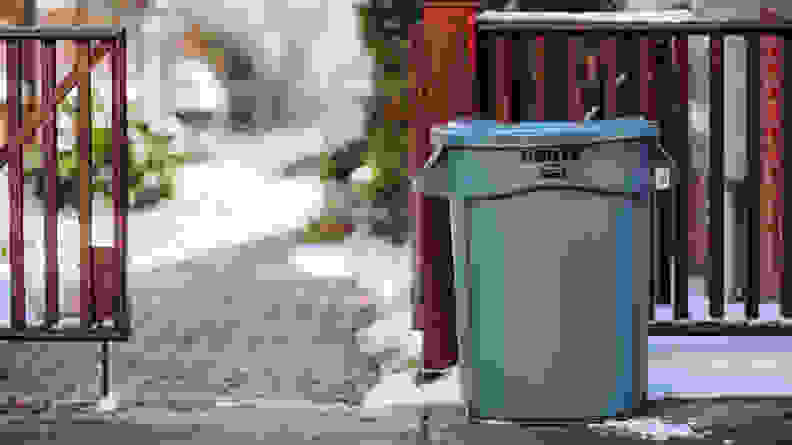 A grey Rubbermaid Brute outdoor trash can on a sidewalk in front of an open fence.