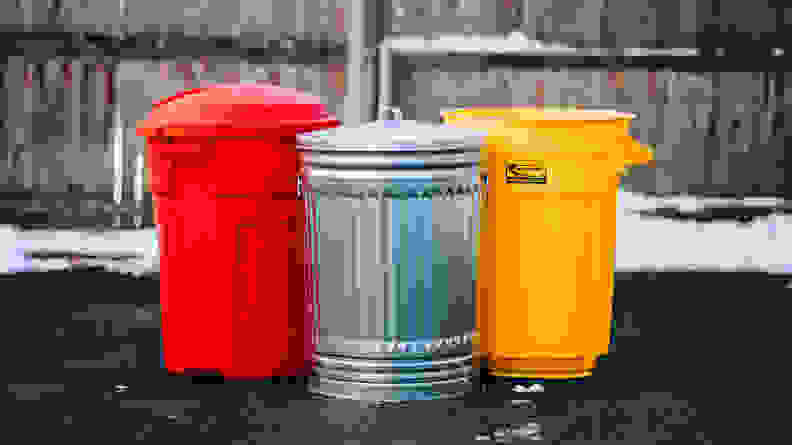 A red trash can, silver steel trash can, and yellow trash can sit on asphalt in front of a wooden fence.