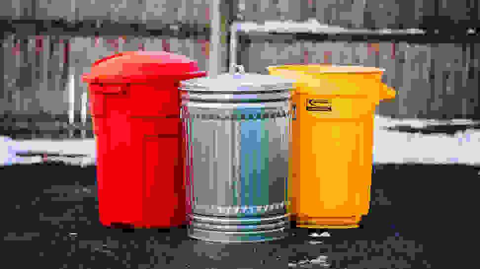 A red Rubbermaid 32 gallon outdoor trash can, Behrens galvanized steel 31 gallon trash can, and a yellow Suncast trash can sitting outside in front of a fence.