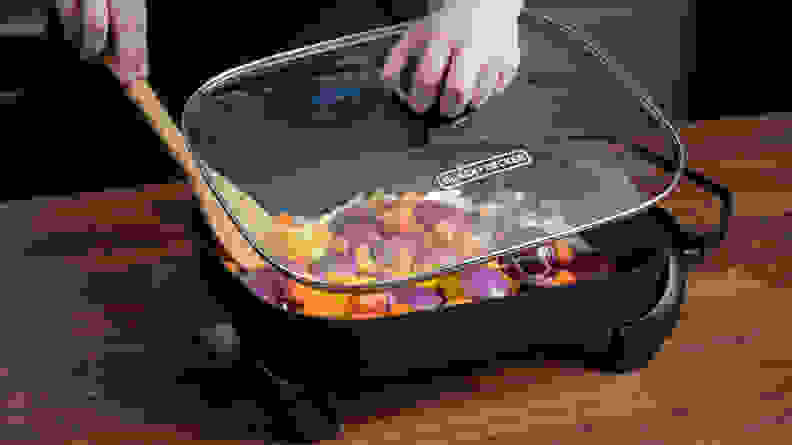 Person removing the lid from Black and Decker electric skillet to stir vegetables with wooden spoon