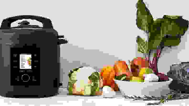 A black pressure cooker sits next to a pile of fresh produce.