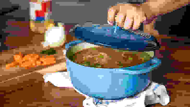 A person lifts the lid of a blue Lodge Dutch oven to reveal chicken soup inside.