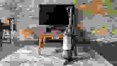 The Dyson Animal 3 in the upright position on blue carpet in front of a television on a wooden stand in front of brick wall.