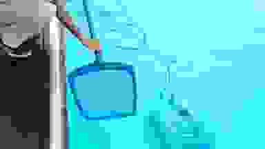 The image shows a pool vacuum cleaner working in the bottom of a pool, while someone uses a net.