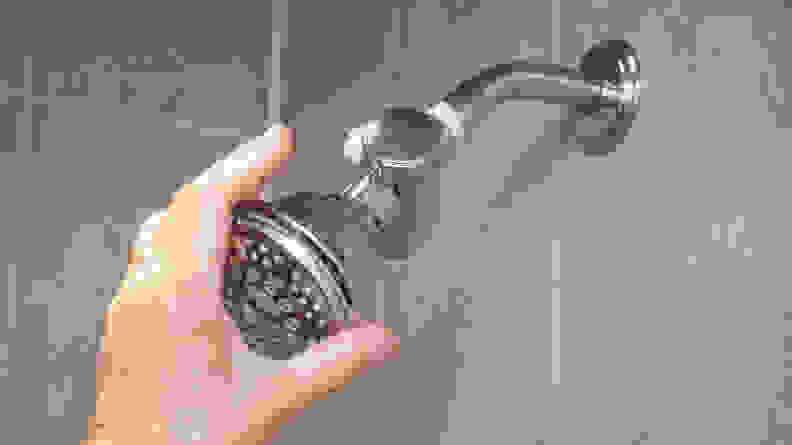 A hand reaches around a silver Hydroluxe showerhead mounted in a shower.