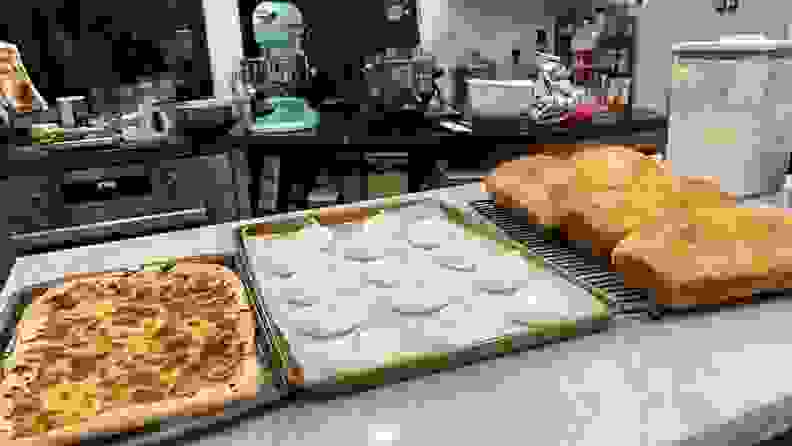 Pizza, meringue cookies and loaves of bread are lined up on a marble table with stand mixers and kitchen equipment in the background.