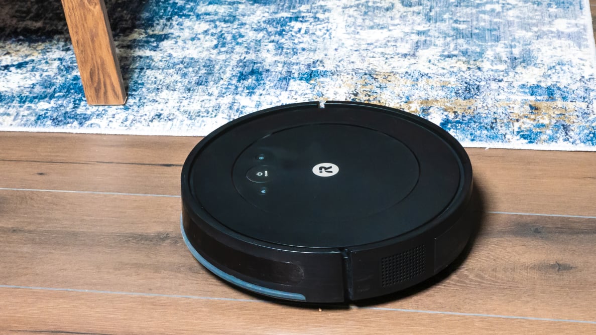 The iRobot Roomba Comobo Essential on a hardwood floor next to carpet.