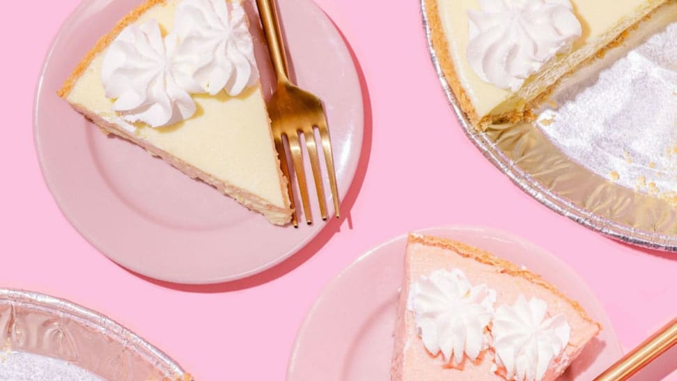 A piece of regular key lime pie and a piece of strawberry key lime pie sit on two pastel pink plates, accompanied by the half-empty aluminum pie tins against a pink background.