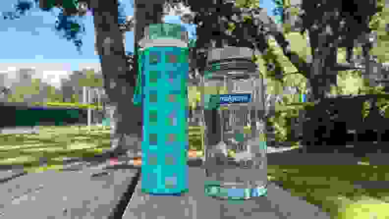 The Ello glass water bottle sits next to the Naglene plastic water bottle on a wooden table at the park with trees in the background