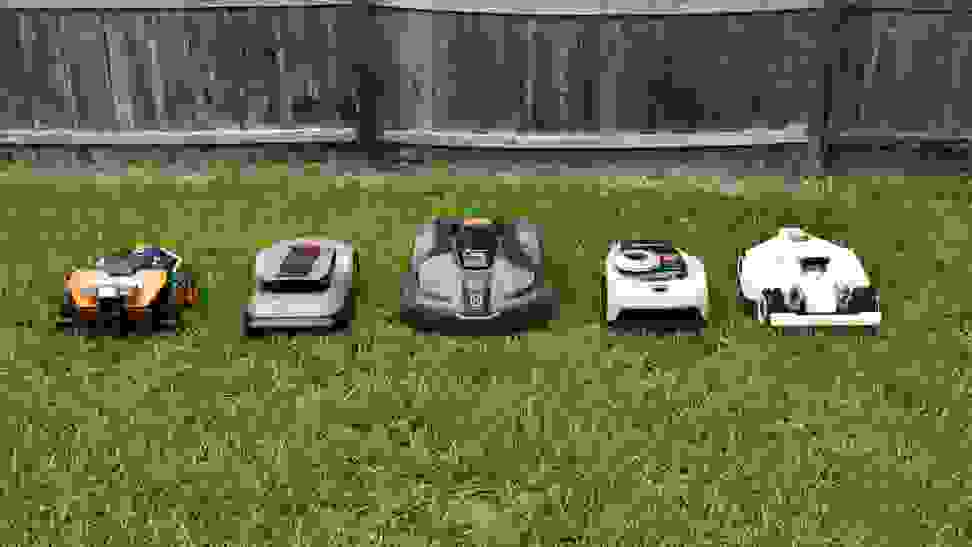 Five robot lawn mowers lined up together in the green grass with a fence behind