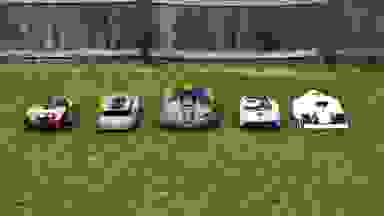 Five robot lawn mowers lined up together in the green grass with a fence behind