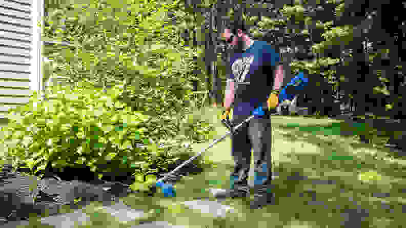 A man uses a blue Dewalt weed wacker to cut grass around the edges of a garden