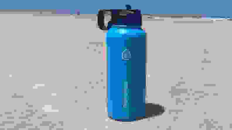 A blue stainless steel water bottle with black lid by ThermoFlask sits on the sand with the ocean in the background