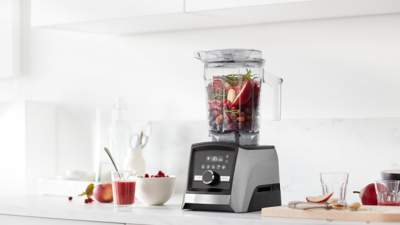 On a counter, there's a Vitamix Ascent Series A3500 blender with grey stainless steel finish. The blending cup is packed with apples and green vegetables, ready to make smoothies.