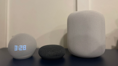 Amazon Echo, Google Home, and Apple HomeKit smart speakers all sit next to each other on a blue table.