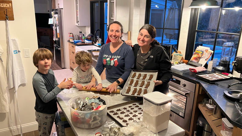 Two women and two children making gingerbread cookies on a table with trays of cutout cookies and a bin of cookie cutters.