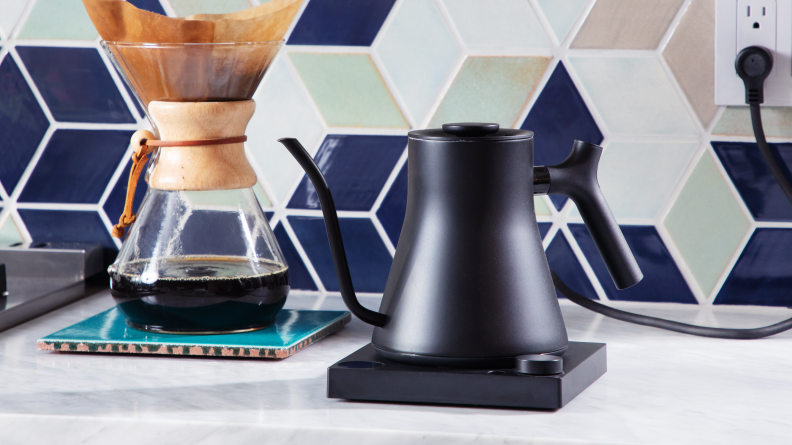 The black Fellow Stagg EKG Pro electric gooseneck kettle next to a Chemex pour-over coffee maker in front of a tile backsplash.