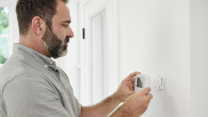 A man with dark hair and a gray shirt stands against a white wall installing a thermostat.