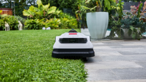 A robotic lawn mower cutting grass precisely along the edge of a neatly maintained lawn, surrounded by lush greenery and a modern patio.