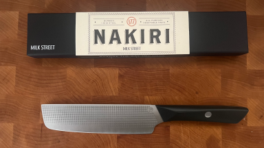 A Milk Street Nakiri Knife underneath the box packaging on top of a wooden cutting board.