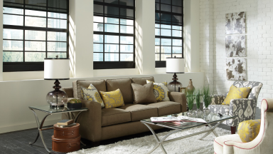 View of a lovely studio apartment living space: a glass coffee table, abstract art on the wall, white walls, black smart blinds, a white decorative lamp, gold throw pillows, and a white rug.