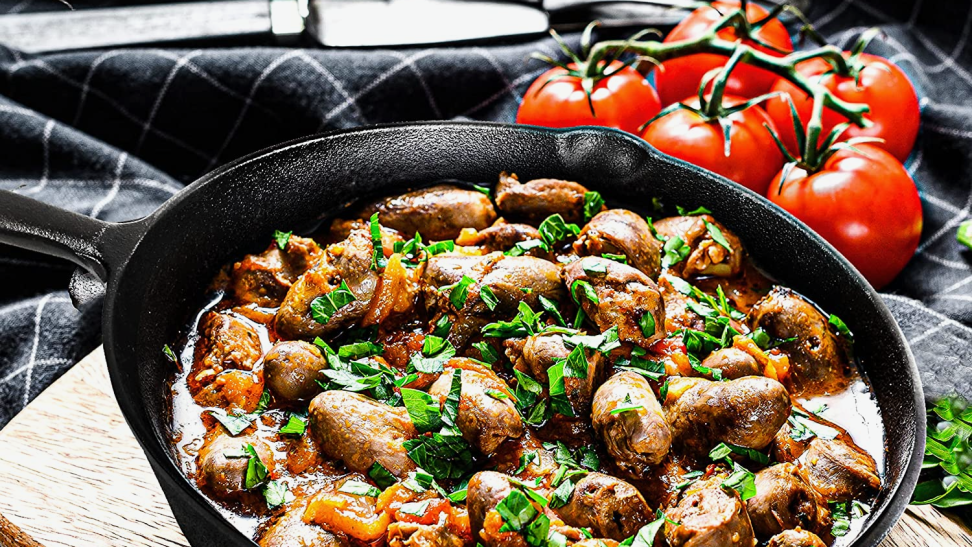 A juicy, savory dish, rich in protein, herbs, and vegetables, sits in a black cast iron skillet. Five ripe tomatoes are on the countertop behind it.